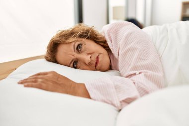 Orta yaşlı beyaz kadın yatak odasında yatakta yatıyor..