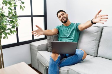 Sakallı genç yakışıklı adam evdeki koltukta oturup kucaklaşmak için kolları açık gülümseyen kameraya bakıyor. Mutluluğu kucaklayan neşeli bir ifade.. 