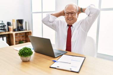 Ofiste çalışan kıdemli bir adam bilgisayarı kullanıyor rahatlıyor ve esniyor, kolları ve elleri başının arkasında mutlu bir şekilde gülümsüyor. 