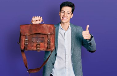 Genç İspanyol adam iş çantasını tutuyor mutlu ve pozitif gülümsüyor, başparmağıyla mükemmel ve onay işareti yapıyor. 