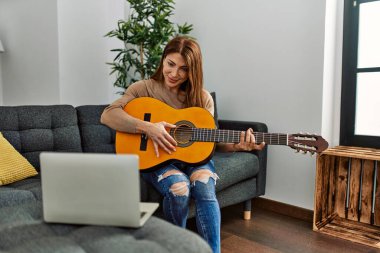 Orta yaşlı beyaz bir kadın online gitar dersi alıyor. Evdeki kanepede oturuyor..