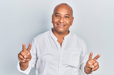 Orta yaşlı, sıradan kıyafetler giyen, gülümseyen, parmaklarını zafer işareti yaparken gösteren Latin bir adam. İki numara.. 