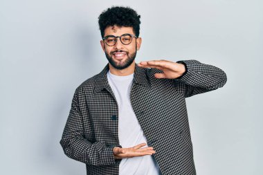 Sakallı, gözlüklü, el kol hareketleri yapan, büyük boy ve ölçü işareti olan genç bir Arap. Kameraya bakıp gülümsüyor. ölçüm kavramı. 
