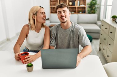 Genç beyaz çift dizüstü bilgisayar kullanıyor ve kahve içiyor. Evdeki masada oturuyorlar..