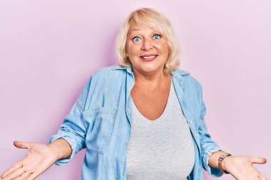 Sıradan kıyafetler giyen orta yaşlı sarışın kadın elleri ve kolları havada, kafası karışık bir ifade takınıyor. şüphe kavramı. 