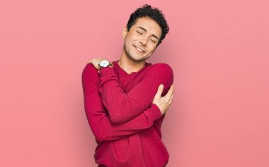 Genç, yakışıklı, günlük kıyafetler giyen, kendine sarılan mutlu ve pozitif, kendinden emin gülümseyen bir adam. Sevgi ve özgüven. 