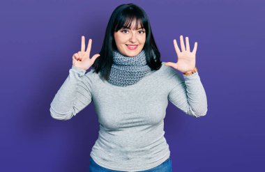 Genç İspanyol asıllı, uzun boylu, kış eşarbı takan, 8 numaralı parmaklarıyla işaret eden, kendinden emin ve mutlu bir şekilde gülümseyen kadın.. 