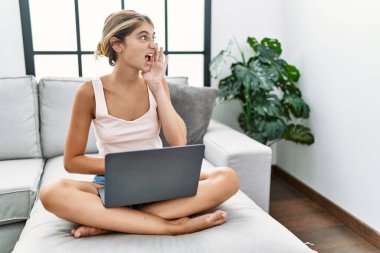 Genç sarışın kadın evinde dizüstü bilgisayar kullanıyor kanepeye oturup bağırıyor ve ağızdan ağıza bağırıyor. iletişim konsepti. 