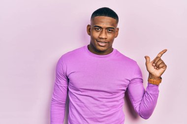Yüzünde kocaman bir gülümseme olan pembe süveter giyen genç siyahi bir adam, kameraya el ile işaret ediyor.. 