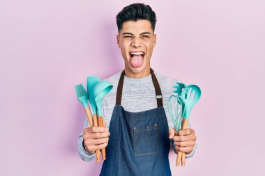 Profesyonel fırıncı önlüğü giyen genç İspanyol adam elinde yemek aletleri, dilini çıkarıp komik bir ifade takınıyor.. 