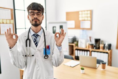 Ofiste sakallı, doktor üniforması giyen ve steteskop takan, gözleri kapalı gülümseyen ve parmaklarıyla meditasyon hareketi yapan İspanyol bir adam. yoga konsepti. 