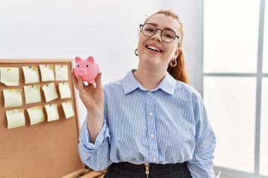 Genç kızıl saçlı kadın domuz kumbarasını ofiste tutuyor. Pozitif ve mutlu görünüyor. Gülümsüyor ve kendine güvenen bir gülümsemeyle dişlerini gösteriyor. 