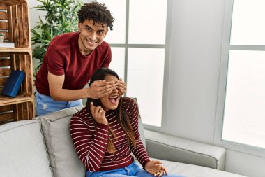 Man surprising his girlfriend with hands on eyes sitting on the sofa at home.