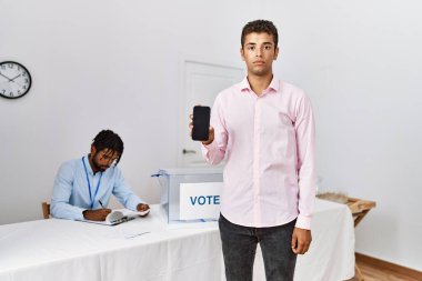 Young hispanic men at political campaign election holding smartphone thinking attitude and sober expression looking self confident  clipart