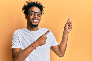 Sakallı, beyaz tişörtlü, gözlüklü, gülümseyen ve kameraya bakan iki eliyle ve parmaklarıyla yüzünü gösteren Afrikalı genç bir Amerikalı.. 
