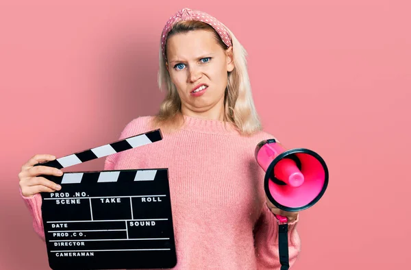 ビデオフィルムのクラップボードとメガホンを持っている若い白人女性は無知で混乱した表情 疑わしい概念 — ストック写真