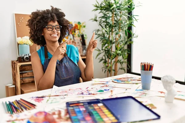 美丽的非洲裔美国女人 在艺术工作室里画着非洲的头发 面带微笑 看着摄像机 用双手和手指指向旁边 — 图库照片
