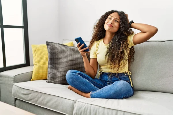 スマートフォンを使って自宅のソファに座っている美しいヒスパニック系の女性が混乱し 疑問に思っています 疑いもなく頭に手を置いて考える 包括的な概念は — ストック写真