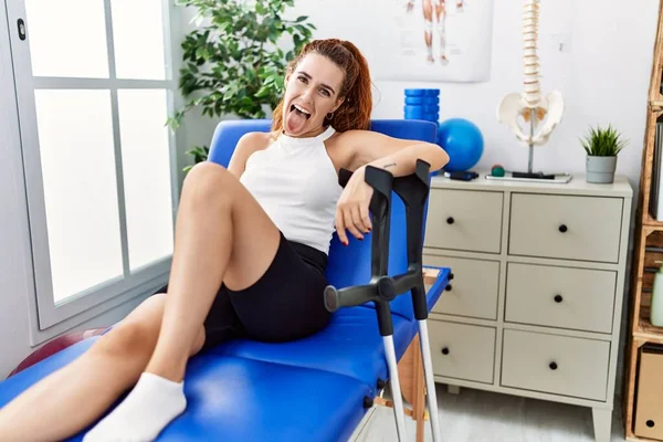 stock image Young redhead woman lying on rehabilitation bed holding crutches sticking tongue out happy with funny expression. emotion concept. 