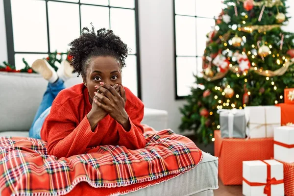 Młoda Afroamerykanka Leżąca Kanapie Koło Choinki Zaszokowana Zakrywaniem Ust Rękami — Zdjęcie stockowe