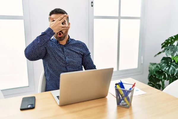 顔と目を手で覆い 恥ずかしい表情で指を通して見て ショックでノートパソコンの目でオフィスで働いて髭を生やした若いヒスパニック系の男 — ストック写真