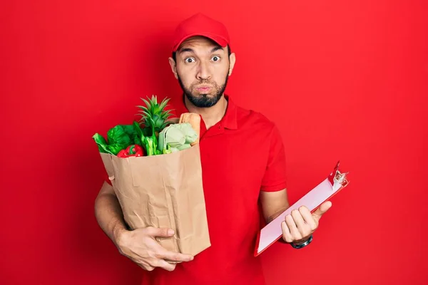一个留着胡子的西班牙男人 身穿信使制服 带着超市的食品杂货和带着滑稽面孔的剪贴板双颊 嘴被空气吹胀 — 图库照片