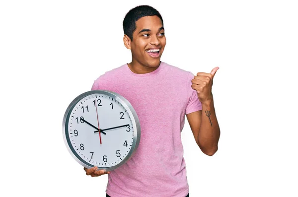 Joven Hombre Hispano Guapo Sosteniendo Gran Reloj Apuntando Con Pulgar —  Fotos de Stock