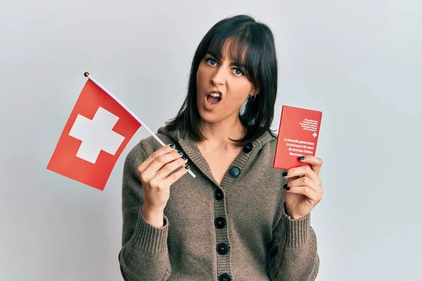 衝撃的な顔でスイスの国旗とパスポートを持っている若いヒスパニック系の女性 懐疑的で皮肉を探して オープン口で驚いて — ストック写真