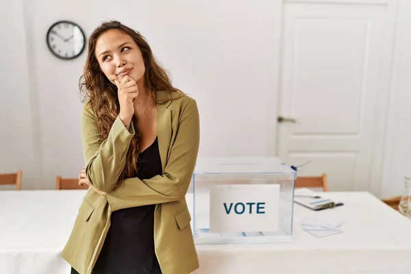 Beautiful hispanic woman standing at political campaign room with hand on chin thinking about question, pensive expression. smiling with thoughtful face. doubt concept.