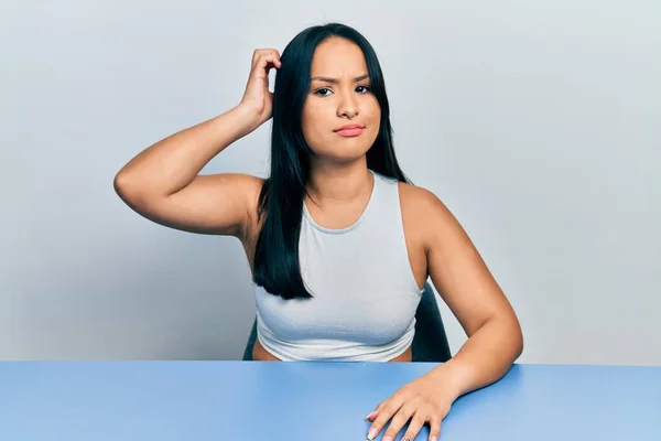 Beautiful hispanic woman with nose piercing sitting on the table confuse and wonder about question. uncertain with doubt, thinking with hand on head. pensive concept.