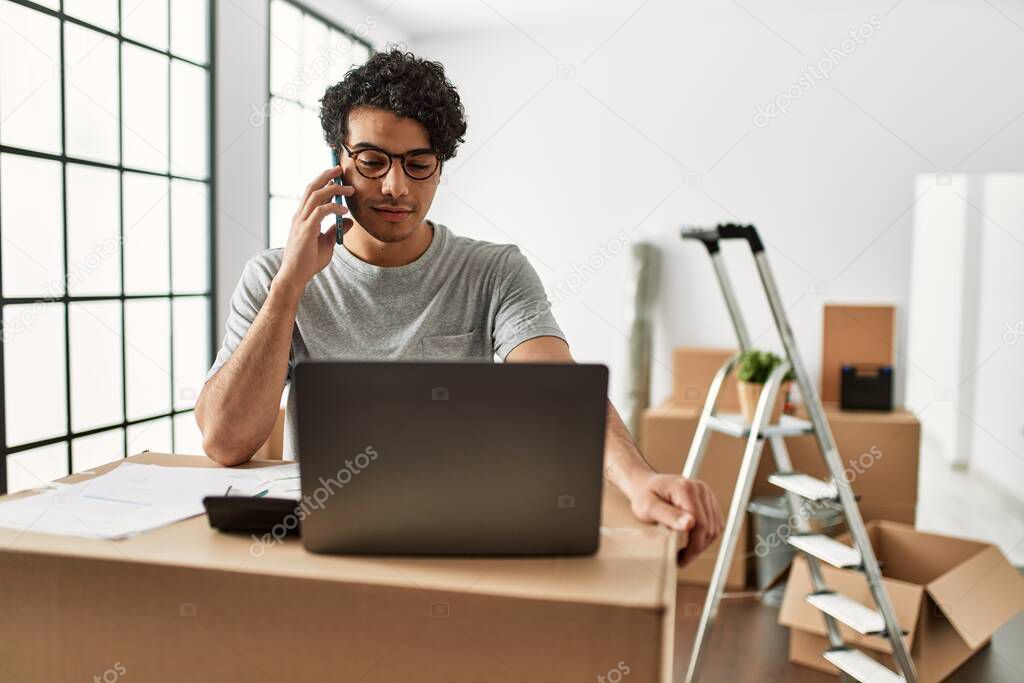 Young hispanic man talking on the smartphone using laptop at new home.