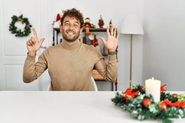 Noel süslemesi masasında oturan sakallı yakışıklı genç adam yedinci parmaklarını gösterip gösterirken kendinden emin ve mutlu bir şekilde gülümsüyor.. 