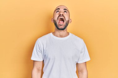 Genç, kel bir adam, günlük beyaz t-shirt giyiyor kızgın ve kızgın bağırıyor sinirli ve öfkeli, öfkeyle bağırıyor. öfke ve agresif konsept. 