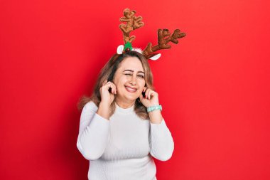 Orta yaşlı, beyaz bir kadın, yüksek sesli müzik sesi için kulaklarını parmaklarıyla kapatan şirin geyik boynuzları takıyor. sağır kavramı. 