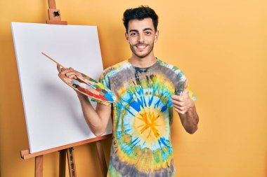 İspanyol asıllı genç adam paletle resim çiziyor, resim sehpasında el hareketleriyle mutlu parmaklar yapıyor. Kameraya bakıp başarıyı gösteren ifade. 
