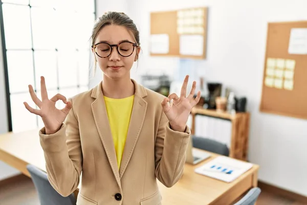 オフィスでビジネススタイルを身に着けている若いブルネットのティーンエイジャーは 指で瞑想のジェスチャーをして閉じ目でリラックスして笑顔 ヨガのコンセプト — ストック写真