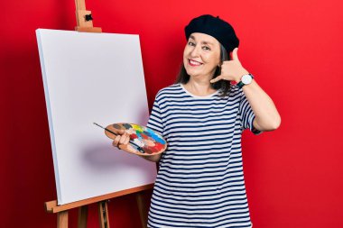 Orta yaşlı İspanyol kadın paletle resim çiziyor resim sehpası ayakta gülümsüyor telefonla el ve parmaklarıyla telefonla konuşuyor gibi yapıyor. iletişim kavramları. 