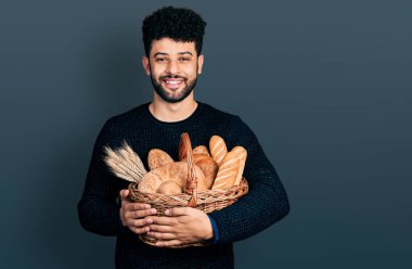 Sakallı genç Arap adam elinde hasırdan sepet tutarken mutlu ve pozitif görünüyor. Kendine güvenen gülümsemesiyle dişlerini gösteriyor. 