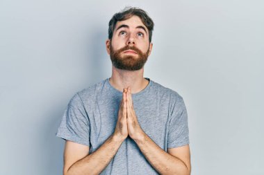 Sakallı, gri tişörtlü beyaz bir adam elleri bağlı yalvarıyor ve dua ediyor. Yüzünde umut ifadesi var. Çok duygusal ve endişeli. Yalvarıyor. 