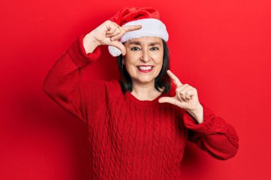 Orta yaşlı İspanyol kadın noel şapkası takıyor gülümsüyor ve mutlu yüzlü eller ve parmaklarla çerçeve yapıyor. yaratıcılık ve fotoğrafçılık kavramı. 