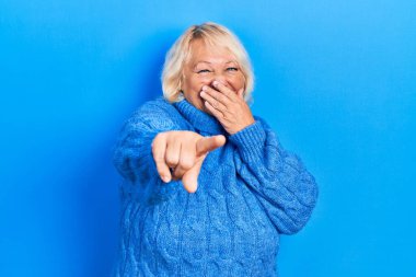 Orta yaşlı, sıradan kıyafetler giyen, sana gülen, eli ağzında, kamerayı gösteren, utanç ifadesi takınan sarışın bir kadın. 