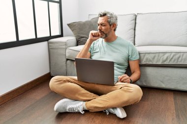 Orta yaşlı İspanyol bir adam oturma odasında dizüstü bilgisayar kullanıyor kendini iyi hissetmiyor ve soğuk algınlığı veya bronşitin belirtisi olarak öksürüyor. sağlık hizmeti kavramı. 