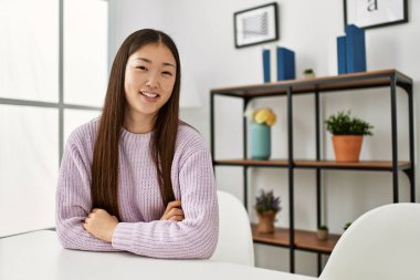 Genç Çinli kız, gündelik giysiler içinde evde masada oturmuş kollarını kavuşturup kameraya bakarak gülümsüyor. pozitif kişi. 