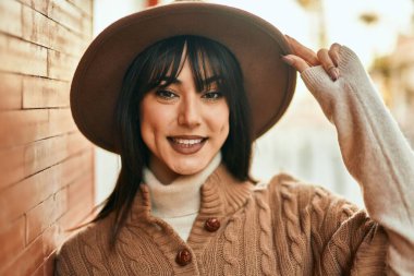 Kış şapkası takan esmer kadın, gülümsüyor tuğla duvara yaslanıyor.