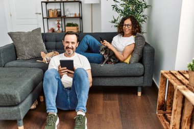 Orta yaşlı İspanyol çift mutlu gülümsüyor ve dokunmatik ped kullanıyor. Evde köpeklerle kanepede oturmak..