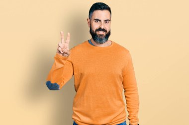 Sakallı, sıradan bir kazak giyen, gülümseyen, parmaklarını zafer işareti yaparken gösteren İspanyol bir adam. İki numara.. 