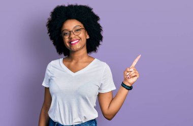 Genç Afro-Amerikalı kadın, günlük beyaz tişörtler giyiyor, yüzünde kocaman bir gülümseme var, kameraya el ile işaret ediyor.. 