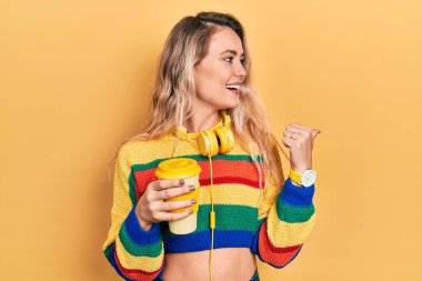 Güzel, genç, sarışın bir kadın, kulaklık takıp kahve içiyor, gülümsüyor, gülümsüyor ve başparmağıyla yan tarafa bakıyor.. 