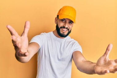 Sakallı, sarı şapkalı genç adam kameraya bakıp sarılmak için kollarını açmış gülümsüyor. Mutluluğu kucaklayan neşeli bir ifade.. 