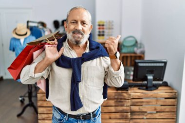 Butik dükkanında alışveriş torbaları tutan yakışıklı bir adam. Arkayı işaret ediyor. El ve baş parmakları havada, kendinden emin gülümsüyor. 